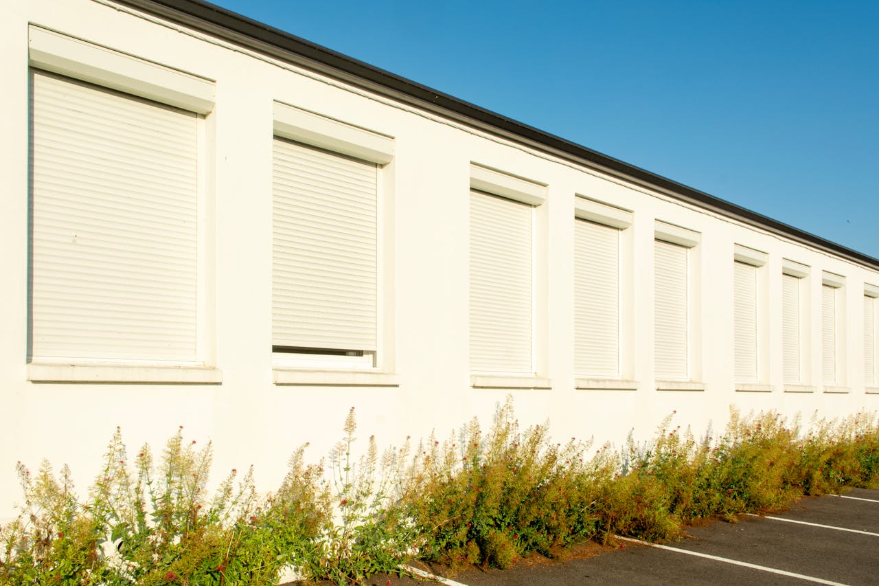 A white building facade with closed shutters and green plants against a clear blue sky.